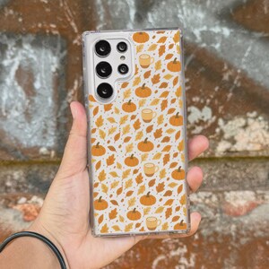 May include: Clear phone case with a fall-themed pattern. The design features orange pumpkins, coffee cups, and autumn leaves on a white background. The case is rectangular and fits a smartphone. The brand name "UBLS" is visible at the bottom.