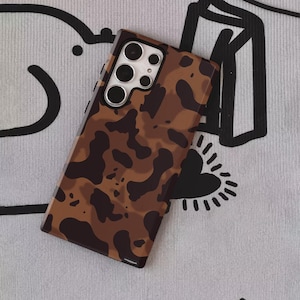 Może przedstawiać: Brązowe i beżowe etui na telefon z wzorem kamuflażu. Etui ma błyszczące wykończenie i wycięcie na aparat z wieloma obiektywami. Etui leży na szarej powierzchni z wzorem. Widoczny jest tekst "UBLS".
