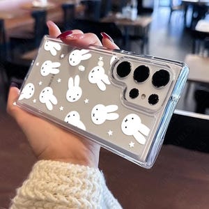 May include: Clear phone case featuring a pattern of white cartoon bunnies and stars on a gray background. The case has a clear border and a camera cutout. The phone case is held in a hand.