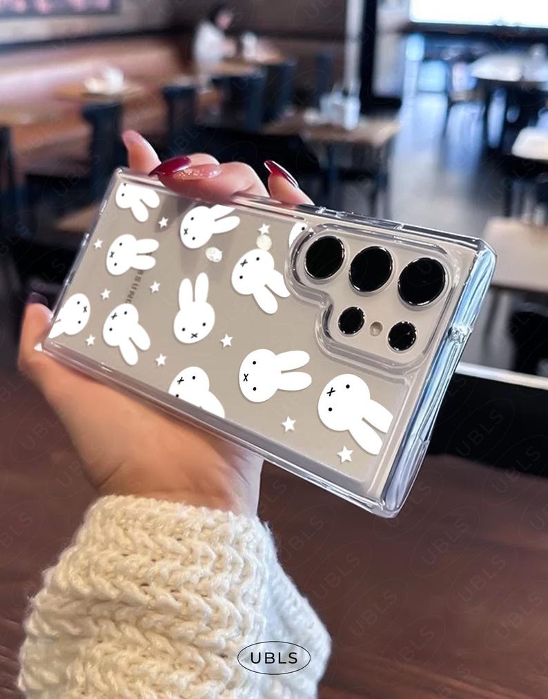 May include: Clear phone case featuring a pattern of white cartoon bunnies and stars on a gray background. The case has a clear border and a camera cutout. The phone case is held in a hand.