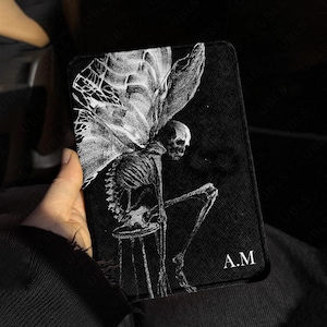 May include: A rectangular black passport holder featuring a white line-art illustration of a winged skeleton sitting on a stool. The design includes the letters A.M in the bottom right corner and a repeating UBLS watermark pattern on the surface.
