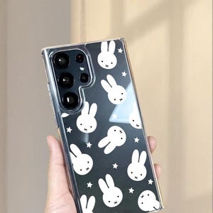 May include: Clear phone case with a black background featuring a pattern of white bunny faces and stars. The case is designed to fit a smartphone and has a clear, transparent appearance. The phone case has a logo that says UBLS.