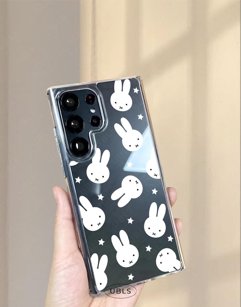 May include: Clear phone case with a black background featuring a pattern of white bunny faces and stars. The case is designed to fit a smartphone and has a clear, transparent appearance. The phone case has a logo that says UBLS.