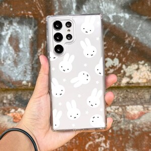 May include: Clear phone case with a repeating pattern of white bunny faces and stars on a light gray background. The case has a clear border and a camera cutout.