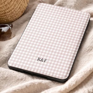 May include: A phone case featuring a light brown and white gingham design. The case displays the initials "R&F". It rests on a beige fabric, with a straw bag and sunglasses visible in the background. The text "UBLS" is also visible.