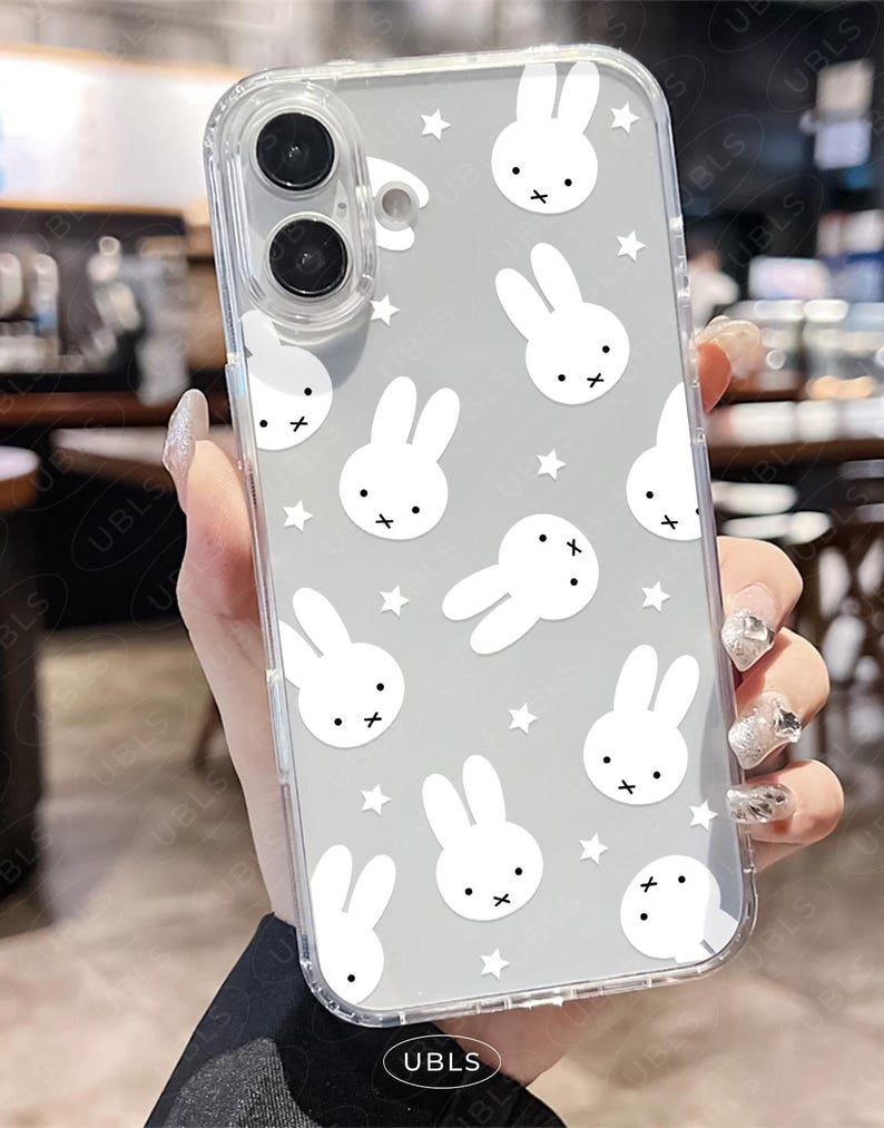 May include: Clear phone case with a gray background featuring white bunny illustrations and small white stars. The case has a transparent border and covers the phone's camera and buttons.