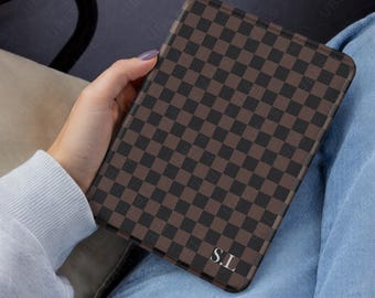 May include: A rectangular electronic reader case featuring a dark brown and black checkered pattern. The bottom right corner of the case displays the white capital letters S.L. The case has rounded corners and a smooth, structured surface.