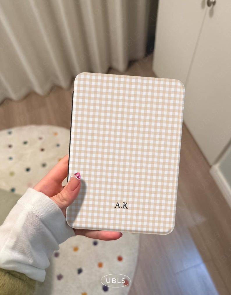 May include: A rectangular phone case with a beige and white gingham pattern. The initials "A.K" are printed in the center. The case is held in a hand.