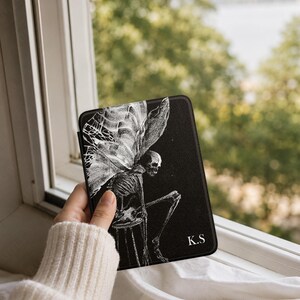 May include: A black e-reader case featuring a white line-art illustration of a winged skeleton sitting on a stool. The design includes the letters K.S in the bottom right corner. The case has a textured surface and a rectangular shape.