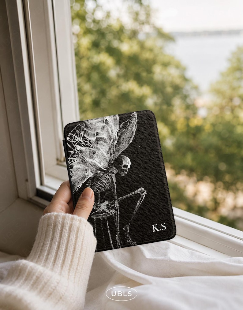 May include: A black e-reader case featuring a white line-art illustration of a winged skeleton sitting on a stool. The design includes the letters K.S in the bottom right corner. The case has a textured surface and a rectangular shape.