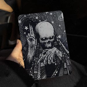 May include: A rectangular e-reader case featuring a high-contrast black and white illustration of a skeletal figure with hollow eyes, wearing a patterned cloak. The figure holds one hand up with two fingers raised against a starry, swirling background.