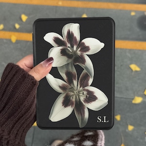 May include: A black e-reader case featuring a graphic print of two white lilies with dark purple centres. The bottom right corner of the case displays the white text S.L. The surface of the case has a textured finish and a rectangular shape.