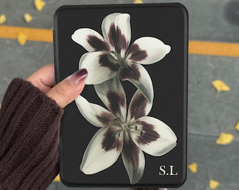 May include: A black e-reader case featuring a graphic print of two white lilies with dark purple centers. The bottom right corner of the case displays the white text S.L. The surface of the case has a textured finish and a rectangular shape.