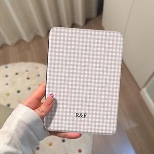 May include: A rectangular phone case featuring a beige and white gingham design. The case has rounded corners and the letters "R&F" are printed on the front. The case is held in a hand.
