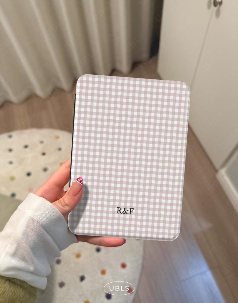 May include: A rectangular phone case featuring a beige and white gingham design. The case has rounded corners and the letters "R&F" are printed on the front. The case is held in a hand.