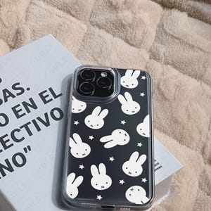 May include: A clear phone case with a black background featuring white bunny faces and stars. The case is on top of a book with the text "AS. EN EL COLECTIVO NO" and the brand name "UBLS".
