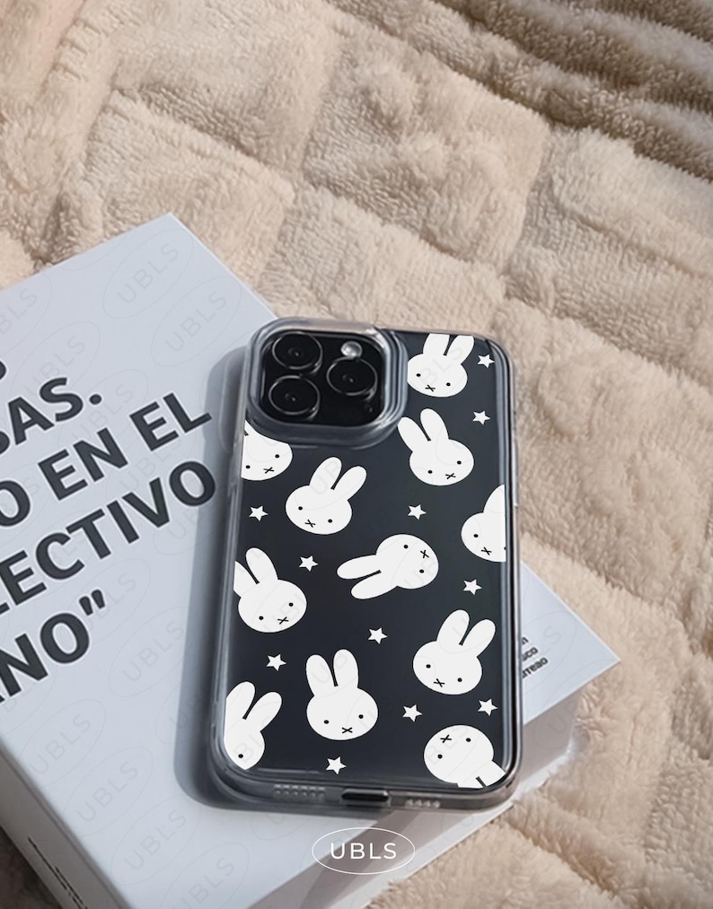 May include: A clear phone case with a black background featuring white bunny faces and stars. The case is on top of a book with the text "AS. EN EL COLECTIVO NO" and the brand name "UBLS".