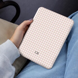 May include: A tablet case featuring a light pink and white gingham design. The initials "CB" are printed on the case. The case is held in a person's hand, with a light blue sweater and jeans visible in the background.