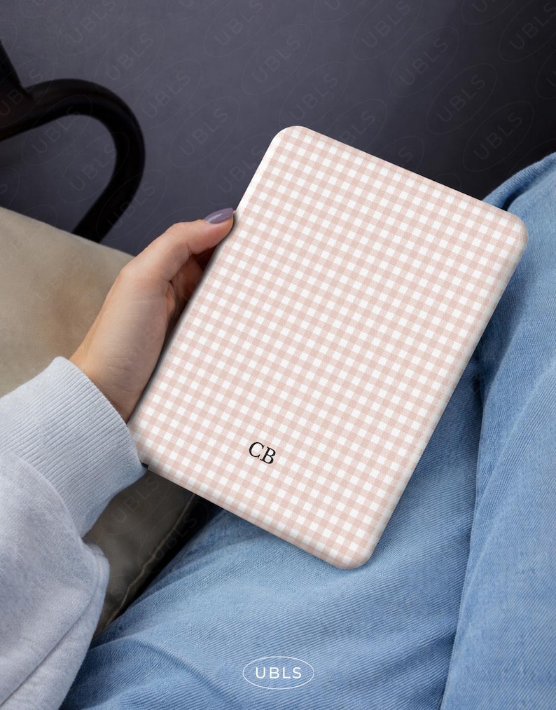 May include: A tablet case featuring a light pink and white gingham design. The initials "CB" are printed on the case. The case is held in a person's hand, with a light blue sweater and jeans visible in the background.
