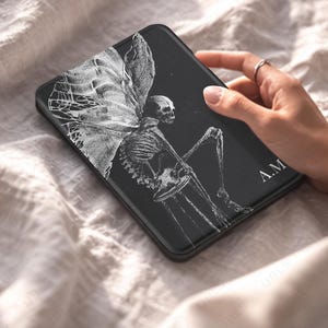 May include: A black rectangular e-reader case featuring a white line art illustration of a skeleton with large, detailed wings sitting on a stool. The initials A.M. are printed in white on the bottom right corner of the cover surface.