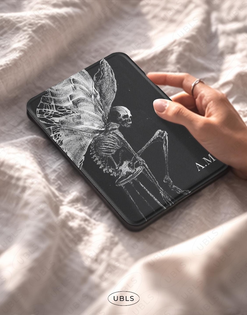 May include: A black rectangular e-reader case featuring a white line art illustration of a skeleton with large, detailed wings sitting on a stool. The initials A.M. are printed in white on the bottom right corner of the cover surface.