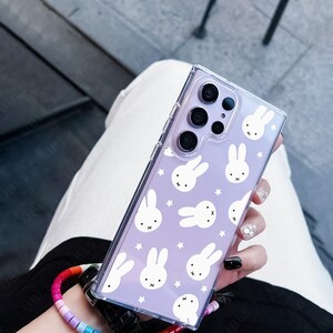 May include: Clear phone case with a lavender background featuring white bunny illustrations and stars. The case is held by a person wearing a rainbow beaded wrist strap. The phone case has a clear border and a camera cutout.