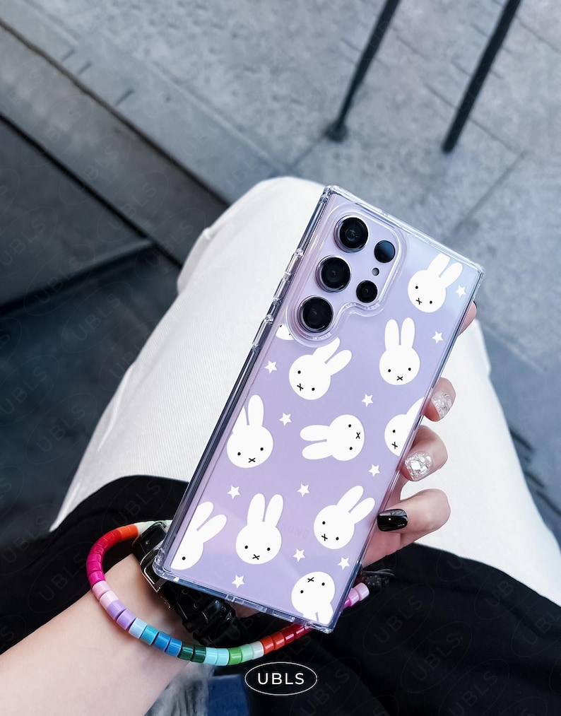 May include: Clear phone case with a lavender background featuring white bunny illustrations and stars. The case is held by a person wearing a rainbow beaded wrist strap. The phone case has a clear border and a camera cutout.