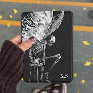 May include: A black rectangular e-reader case featuring a white line-art illustration of a winged skeleton sitting on a stool. The design includes the white text K.S in the bottom right corner. The case has a textured surface and rounded corners.