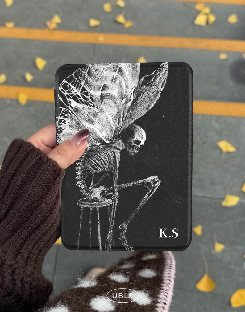 May include: A black rectangular e-reader case featuring a white line-art illustration of a winged skeleton sitting on a stool. The design includes the white text K.S in the bottom right corner. The case has a textured surface and rounded corners.