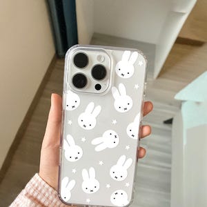 May include: Clear phone case with a repeating pattern of white bunny faces and stars on a gray background. The case has a clear border and covers the back and sides of the phone. The phone's camera lenses are visible through the case.