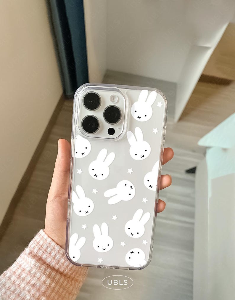 May include: Clear phone case with a repeating pattern of white bunny faces and stars on a gray background. The case has a clear border and covers the back and sides of the phone. The phone's camera lenses are visible through the case.