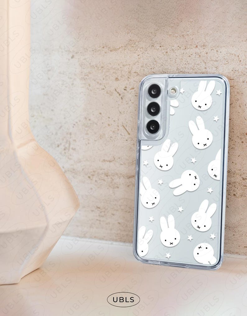 May include: Clear phone case featuring a repeating pattern of white cartoon bunny faces and stars. The case has a transparent border and is designed to fit a smartphone. The UBLS logo is visible at the bottom.