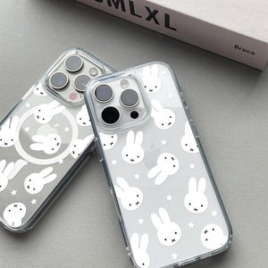 May include: Two clear phone cases with a white bunny and star pattern. The cases have a transparent design, allowing the phone's color to show through. The cases are on a gray surface.