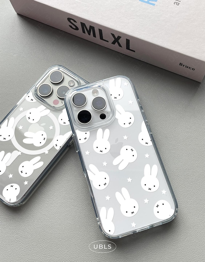 May include: Two clear phone cases with a white bunny and star pattern. The cases have a transparent design, allowing the phone's color to show through. The cases are on a gray surface.