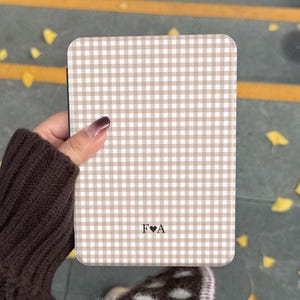 May include: A rectangular phone case with a tan and white gingham pattern. The case has rounded corners and features the initials "F&hearts;A" at the bottom. The case is held in a person's hand.