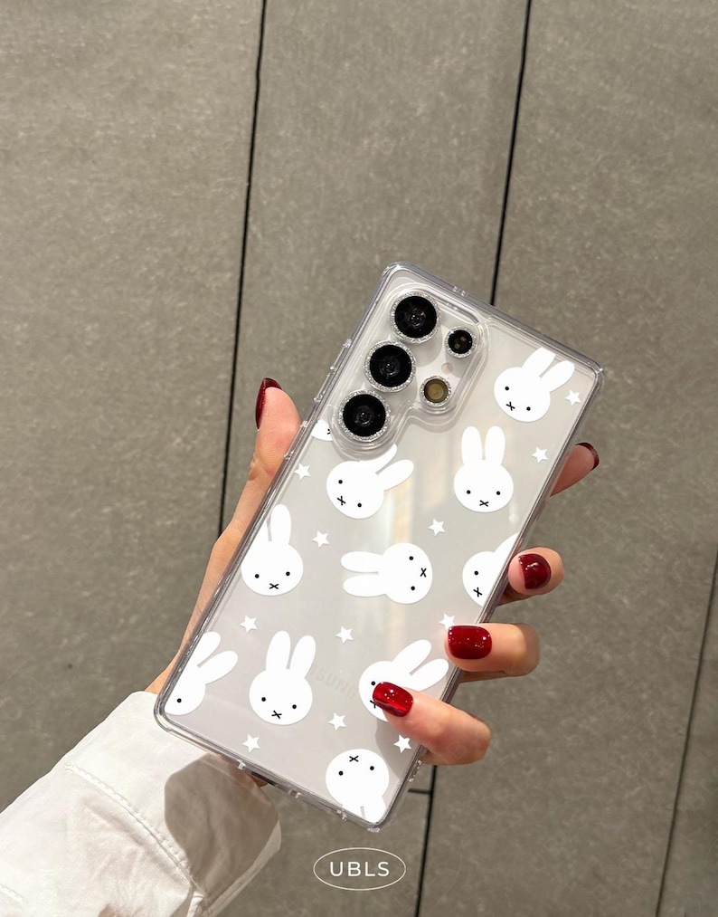 May include: Clear phone case with a white bunny and star pattern. The case has a clear border and covers the back of a smartphone. The text "UBLS" is visible at the bottom of the case.