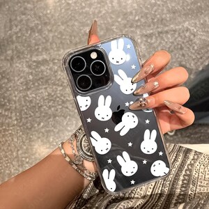 May include: Clear phone case with a black and white bunny pattern. The case has a transparent border and covers the phone's back and sides. The design features white bunny faces and stars on a black background.