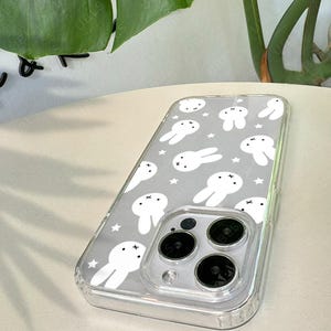 May include: Clear phone case with a gray background featuring a pattern of white bunny illustrations and stars. The case has a clear bumper and covers the phone's camera lenses. The text "UBLS" is visible.