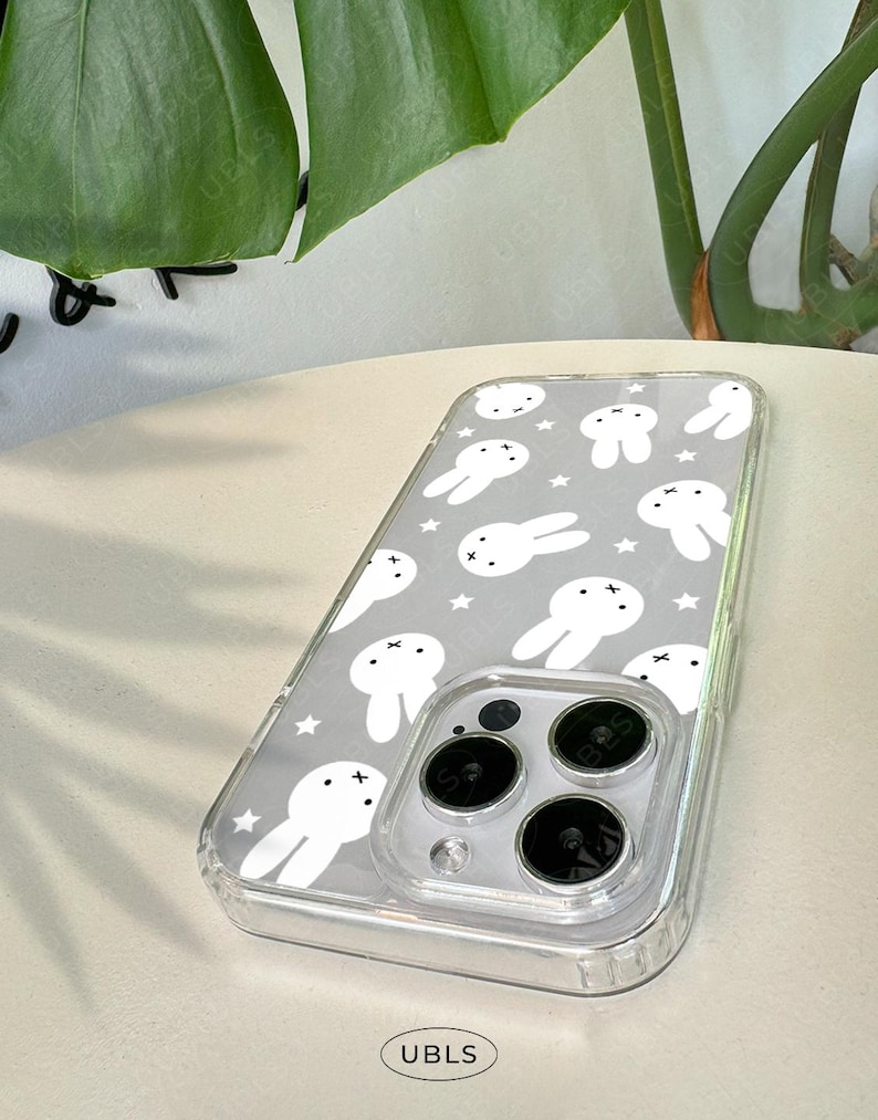 May include: Clear phone case with a gray background featuring a pattern of white bunny illustrations and stars. The case has a clear bumper and covers the phone's camera lenses. The text "UBLS" is visible.