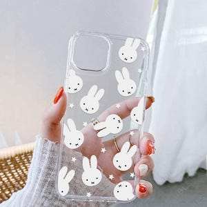 May include: Clear phone case featuring a repeating pattern of white cartoon bunny faces and stars. The case has a cutout for the camera and is held in a hand with painted nails. The brand name "UBLS" is printed at the bottom.