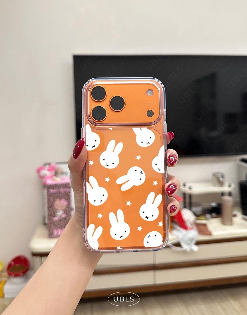 May include: Clear phone case with an orange back featuring a pattern of white bunny faces and stars. The case has a clear border and a camera cutout. The brand name "UBLS" is printed at the bottom.