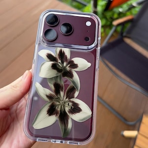 May include: A clear smartphone case featuring a design of two white lilies with dark maroon centers, placed over a deep maroon phone back. The case has a raised camera cutout section and a transparent border, with the text UBLS printed at the bottom.