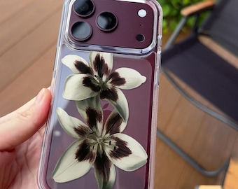 May include: A clear smartphone case featuring a design of two white lilies with dark maroon centres, placed over a deep maroon phone back. The case has a raised camera cutout section and a transparent border, with the text UBLS printed at the bottom.
