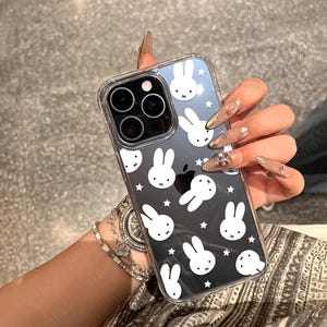 May include: Clear phone case with a black and white bunny pattern. The case has a clear border and covers the back of a smartphone. The design features multiple bunny faces and stars.