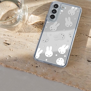 May include: Clear phone case with a white bunny and star pattern. The case is on a light brown surface. The phone case has a clear border and a camera cutout.