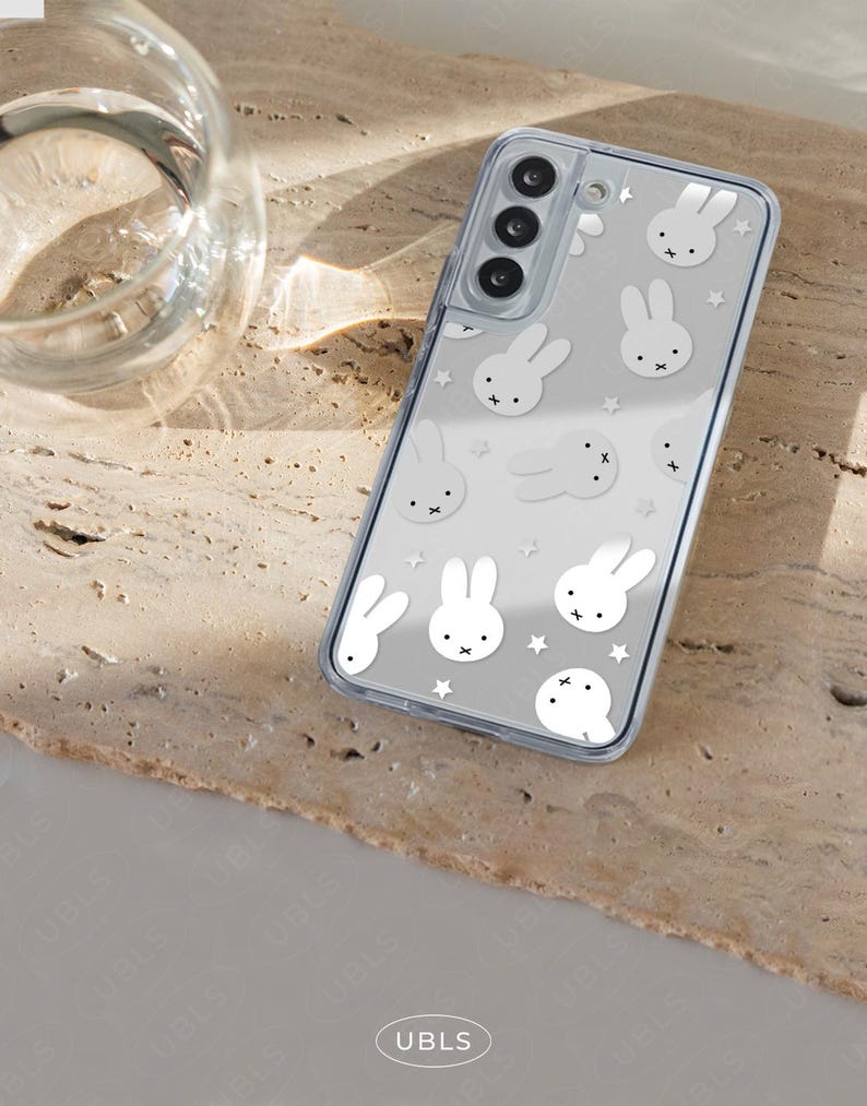 May include: Clear phone case with a white bunny and star pattern. The case is on a light brown surface. The phone case has a clear border and a camera cutout.