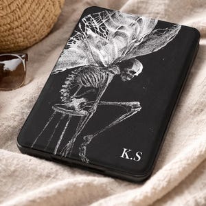 May include: A black rectangular e-reader case featuring a white line art illustration of a winged skeleton sitting on a stool. The bottom right corner of the case displays the white capital letters K.S. The case has a smooth, matte finish.