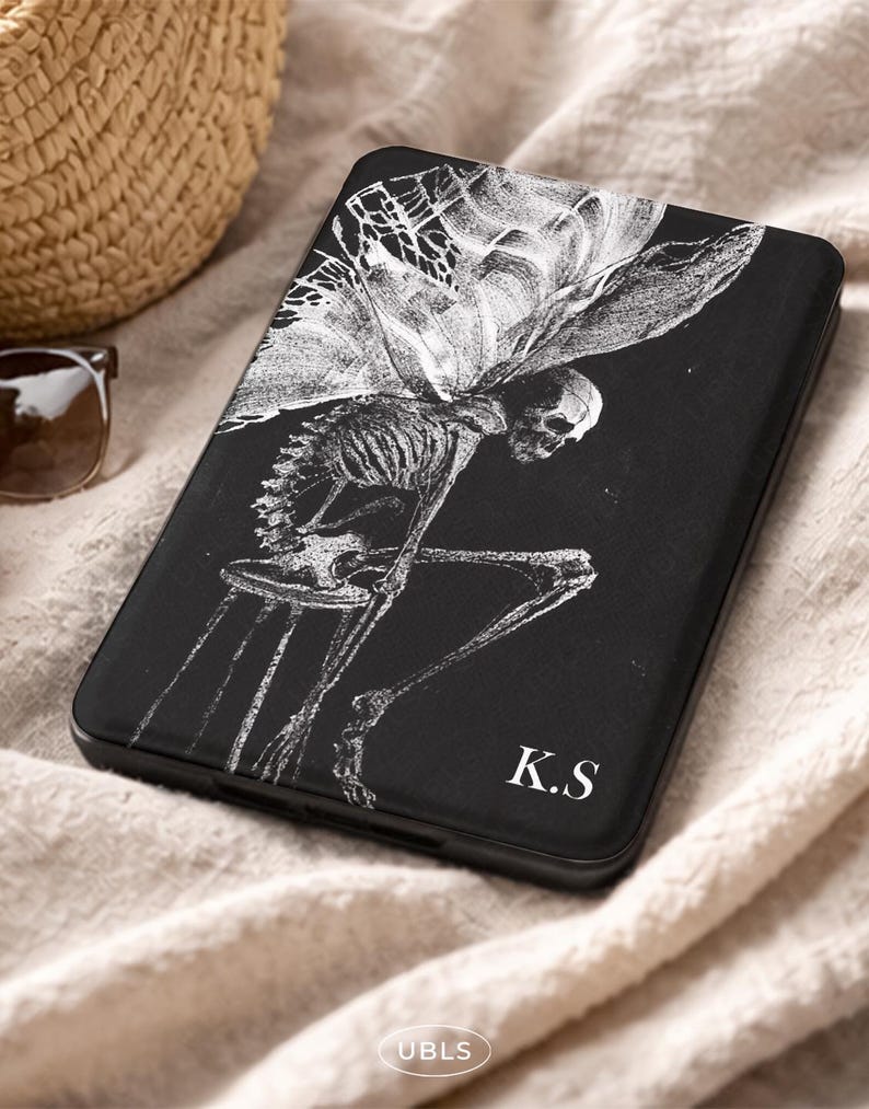 May include: A black rectangular e-reader case featuring a white line art illustration of a winged skeleton sitting on a stool. The bottom right corner of the case displays the white capital letters K.S. The case has a smooth, matte finish.