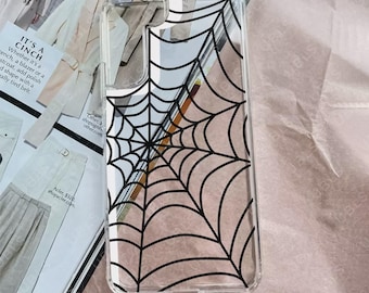 May include: A clear rectangular phone case featuring a black spider web graphic printed across the back. The case has a cutout for a camera lens in the upper left corner and a transparent border. The design consists of thin black lines.