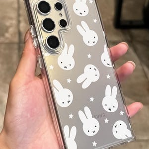 May include: Clear phone case with a white bunny and star pattern. The case has a clear border and covers the back of the phone. The phone has multiple camera lenses.